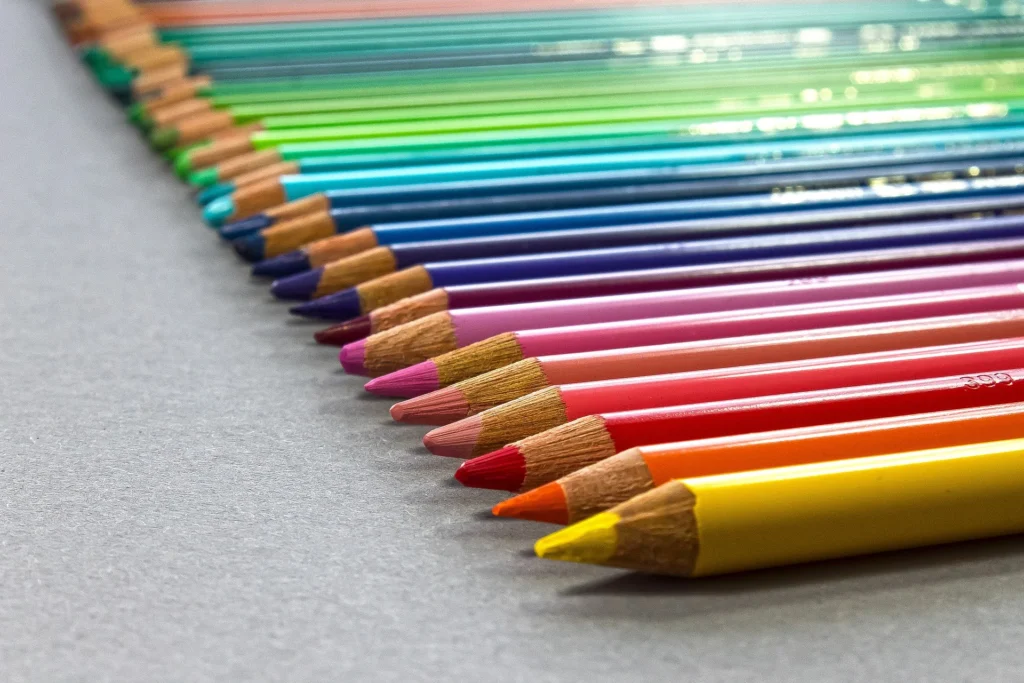 Buntstifte in allen Farben des Regenbogens, schräg nebeneinander auf grauem Papier aufgereiht