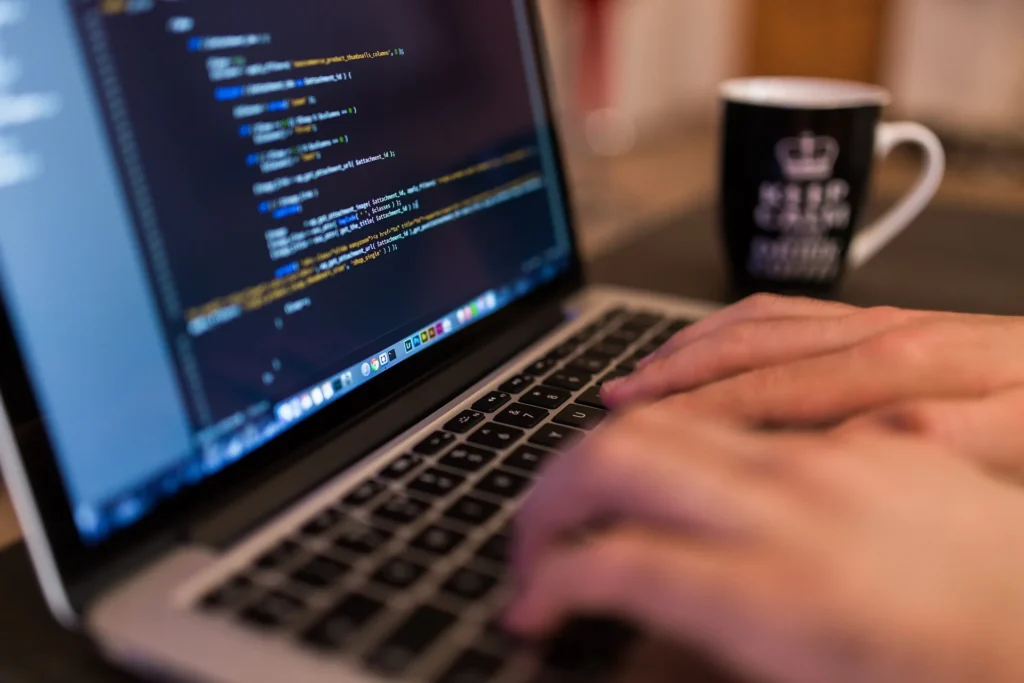 Hände tippen auf einem Laptop, auf dem Bildschirm farbiger Programmcode, im Hintergrund eine Tasse